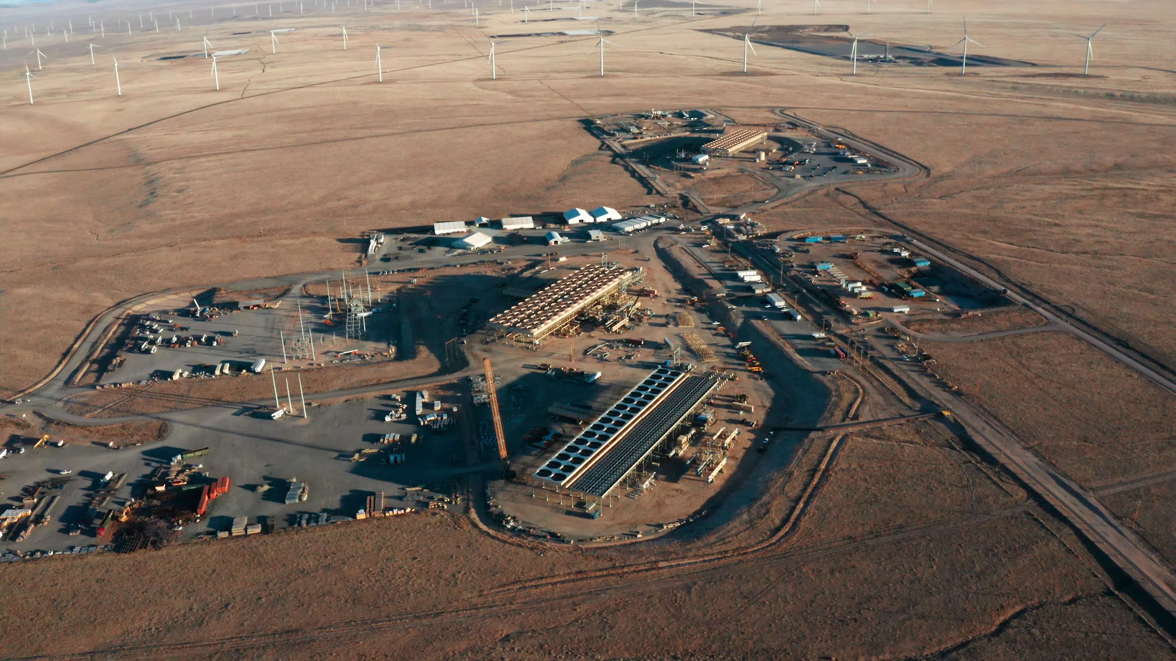 Top-down aerial of Fervo Energy's Cape Station power block showing parallel cooling arrays, transformer yard, and a wind farm on the horizon by Motion Giraffx.