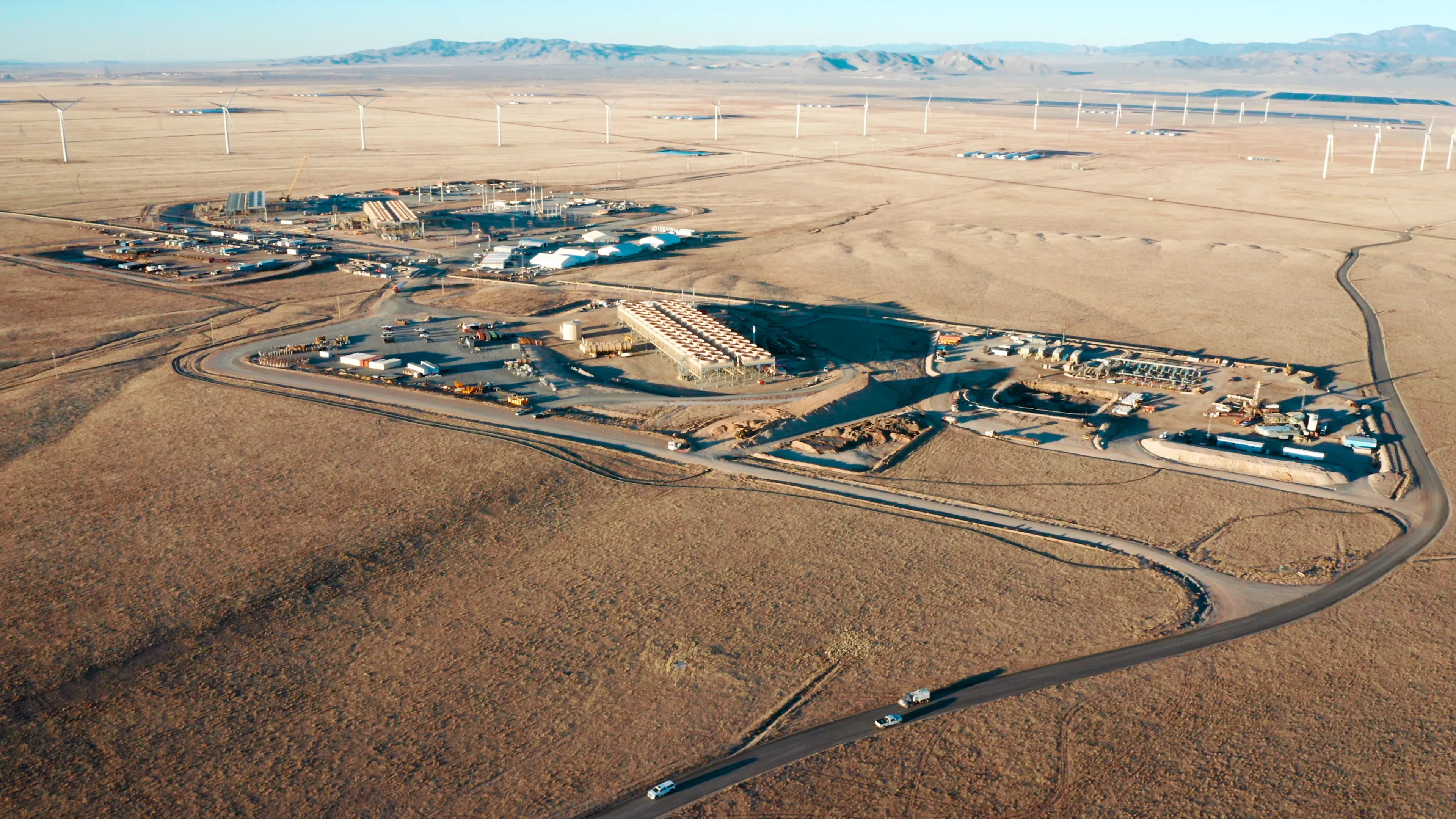 Aerial of service trucks driving in to the Fervo Cape Station power block with the wind farm and mountains behind by Motion Giraffx.