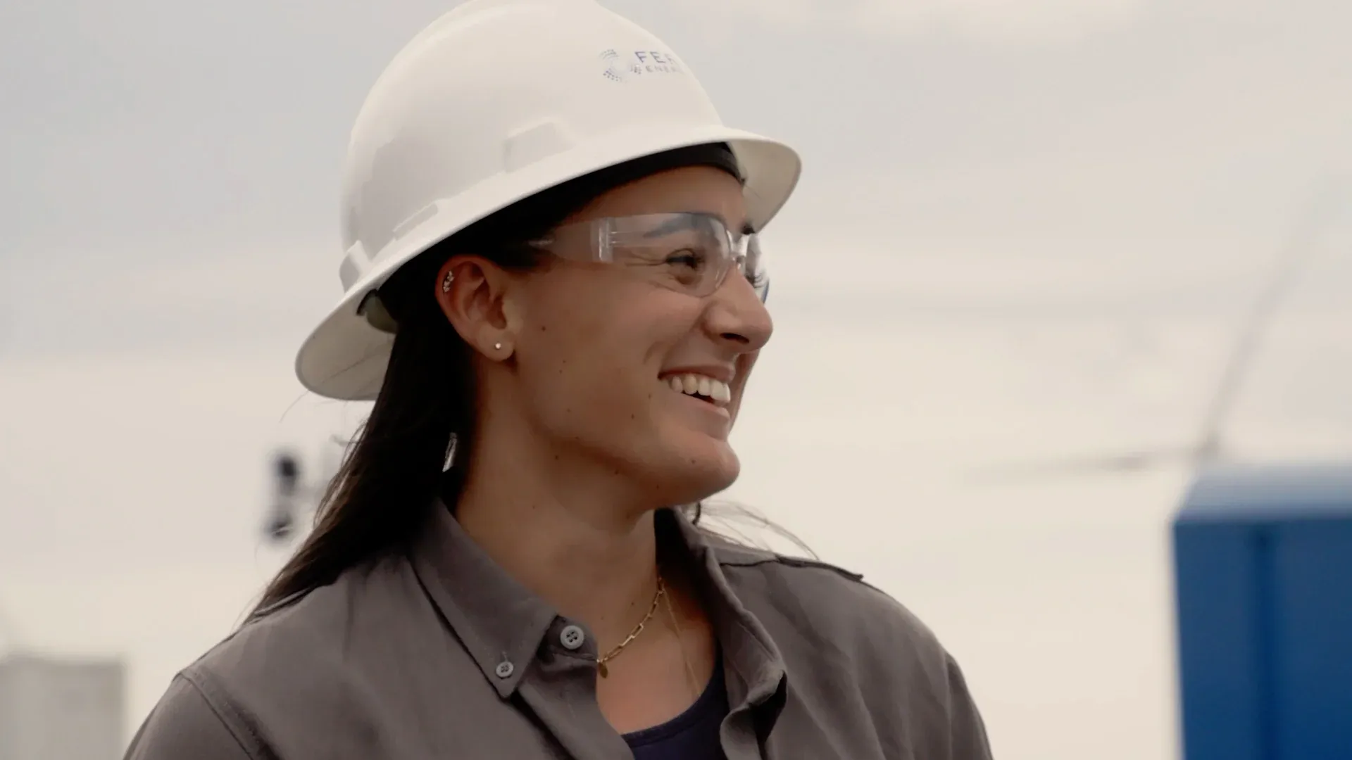 Cinematic profile of a Fervo female engineer in a branded hard hat with shallow depth-of-field and wind turbine bokeh background, from the roadshow film by Motion Giraffx.