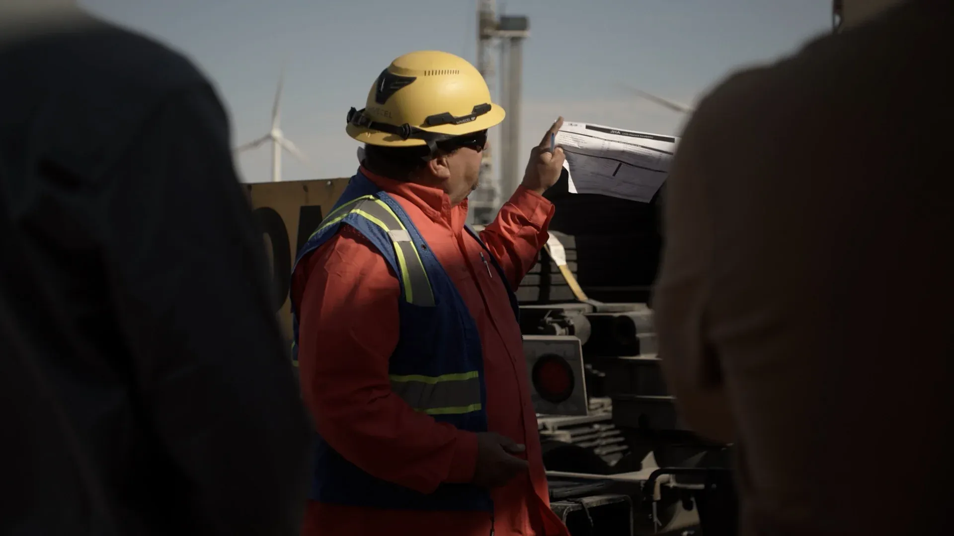 Fervo field technician in FR coveralls and hard hat leading a job hazard analysis safety briefing at a drill site, captured by Motion Giraffx.